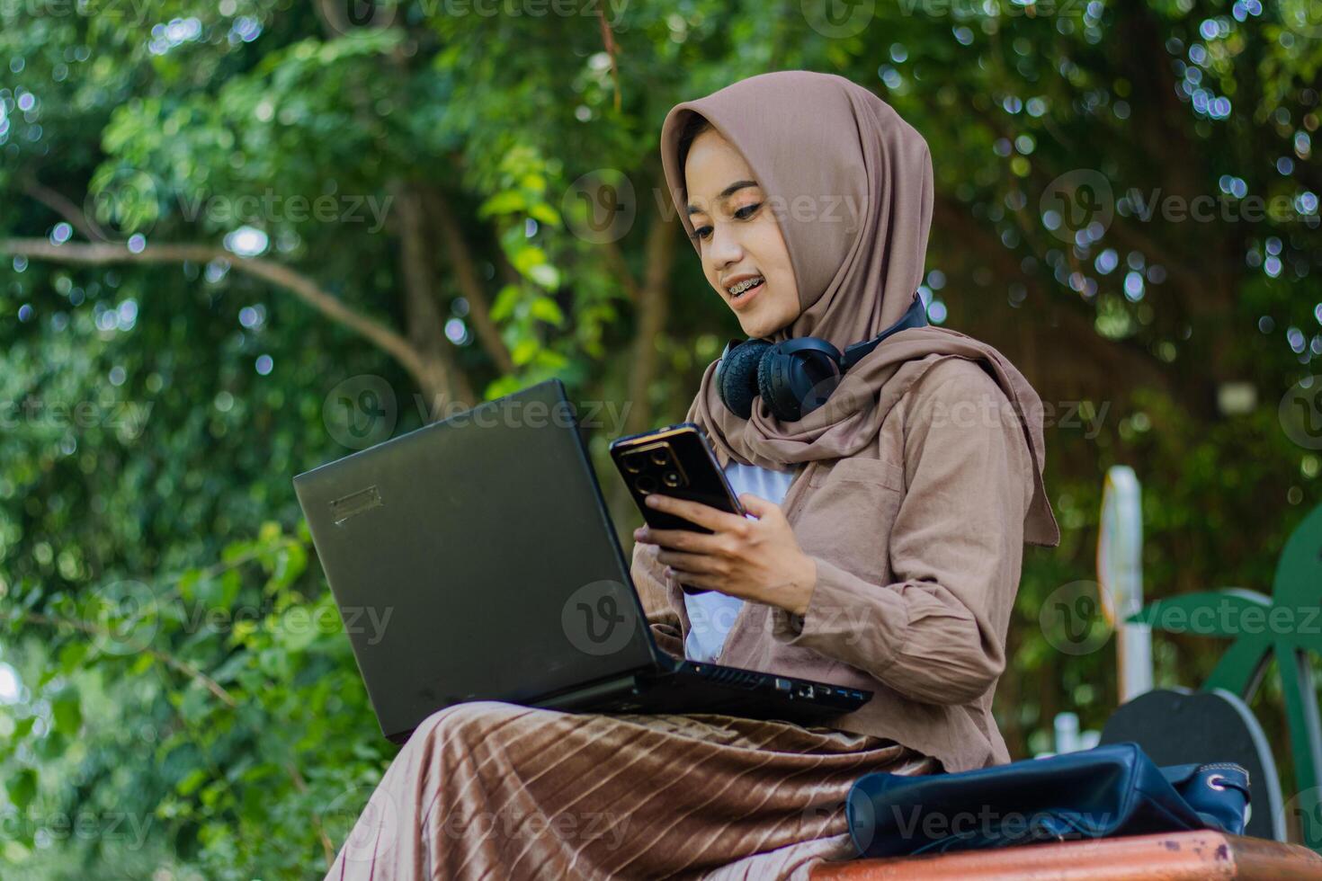 Portrait of a cheerful Asian hijab woman holding a smartphone gadget while operating a laptop sitting in an outdoor park. female student for education, technology and lifestyle concept photo