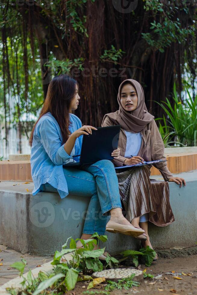 A pair of Asian women candidly discuss studying together carrying laptops, books and clipboards in the campus park. female students for education, promotion and lifestyle concepts photo
