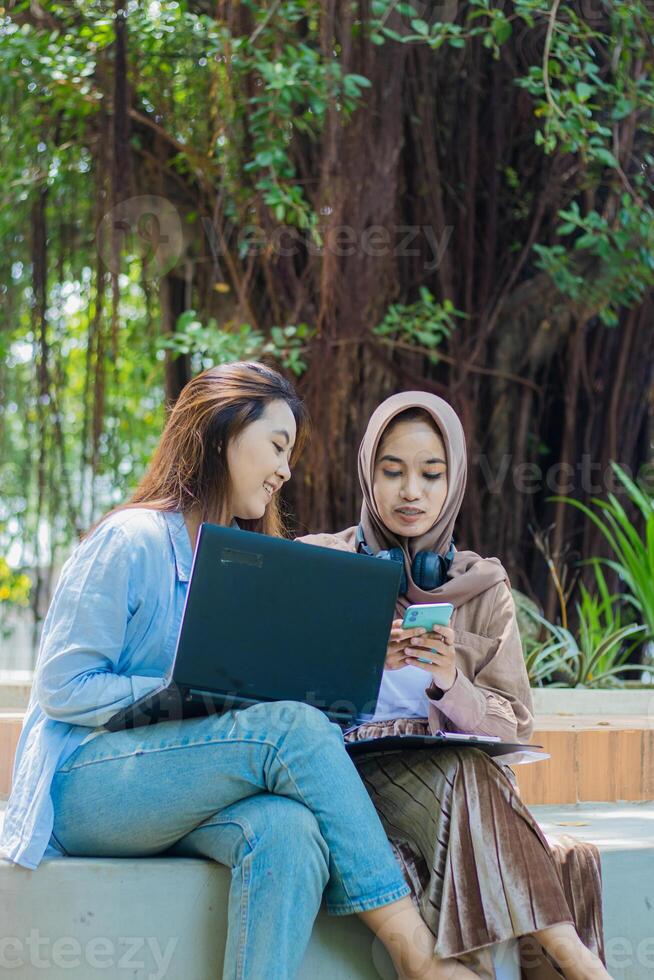 A pair of serious indonesia women typing an online shopping order on a smartphone gadget while holding a laptop, books and clipboard in the campus park. students for education and lifestyle concepts photo