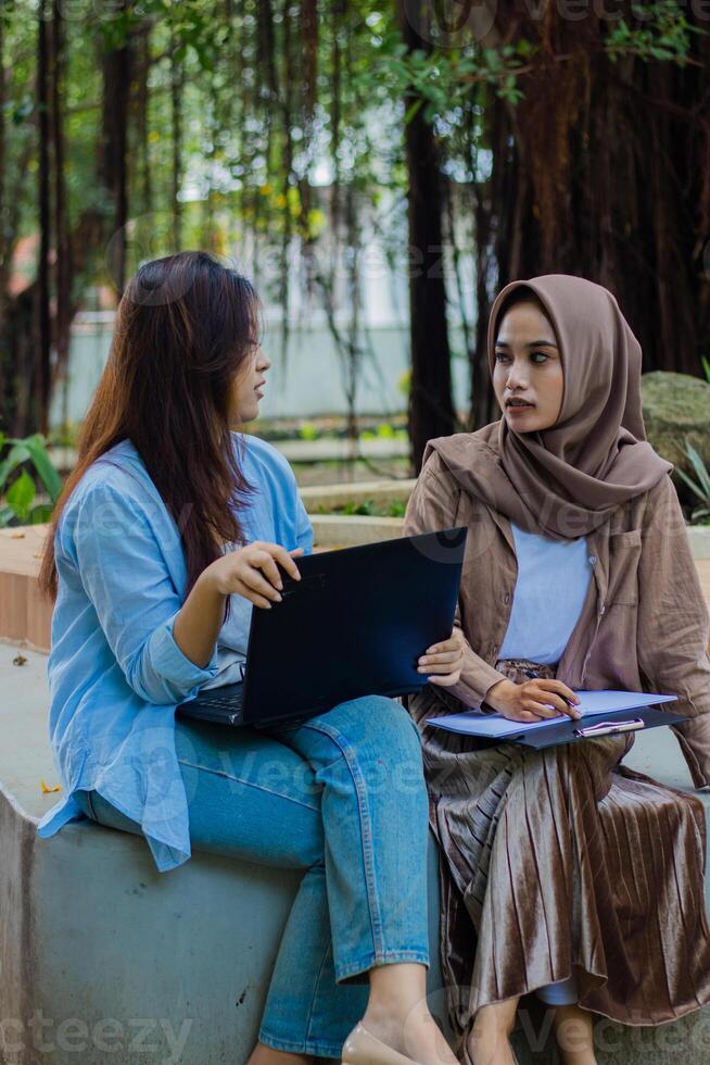 A pair of indonesia women candidly discussing school assignments carrying laptops, books and clipboards in the campus park. female students for education, promotion and lifestyle concepts photo