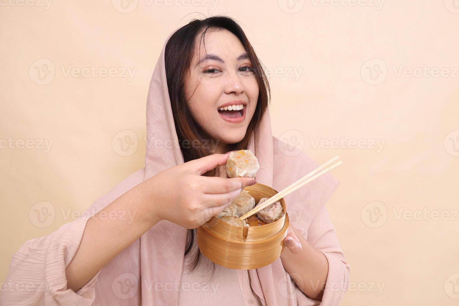 Asian woman wearing a hijab is very happy holding dimsum and a wooden bowl in front of her containing chopsticks and delicious and delicious dimsum photo