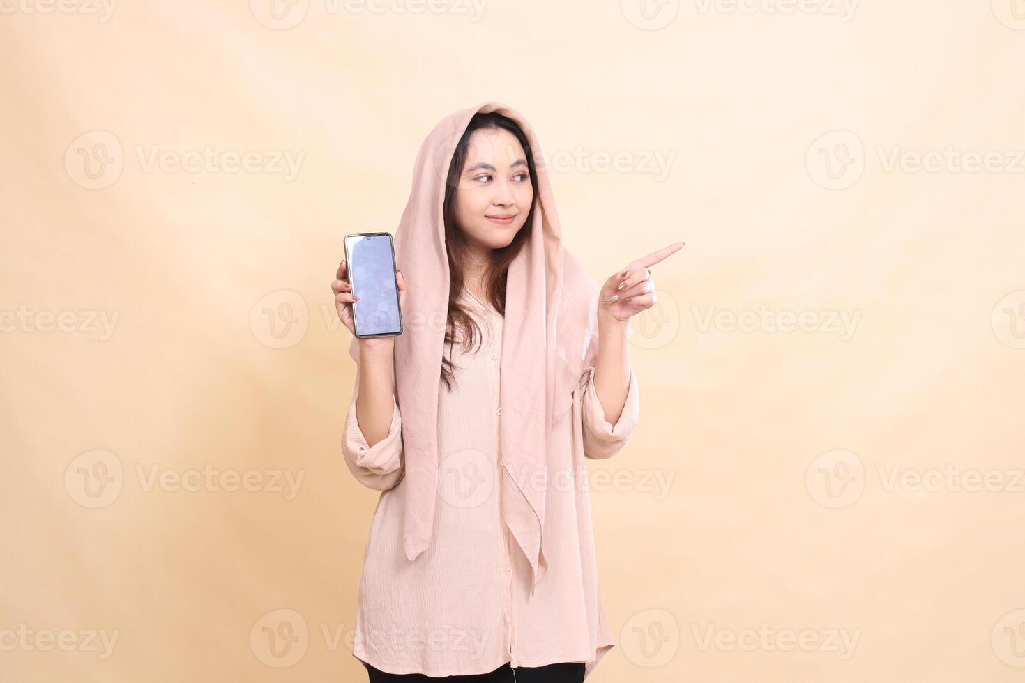 indonesia woman smiles broadly to the left with her left pointing hand and holds the cellphone gadget screen. young beautiful for business, technology, fashion photo