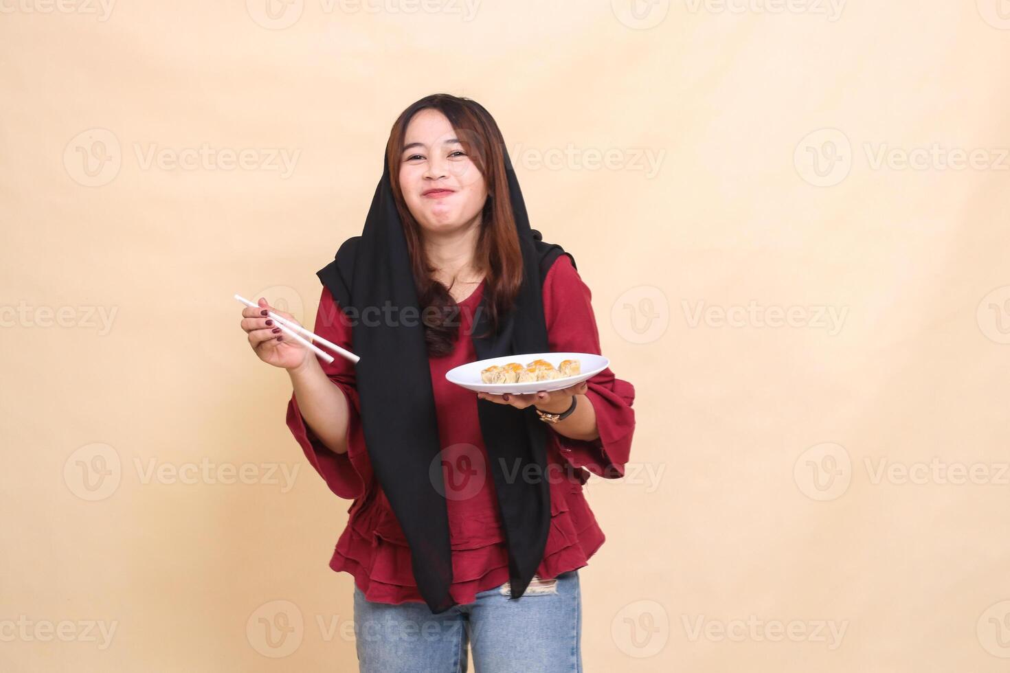 beautiful mature Asian woman wearing a red hijab, smiling, eating dimsum using chopsticks and carrying a plate containing dimsum Chinese food candid. used for food, health and culinary content photo