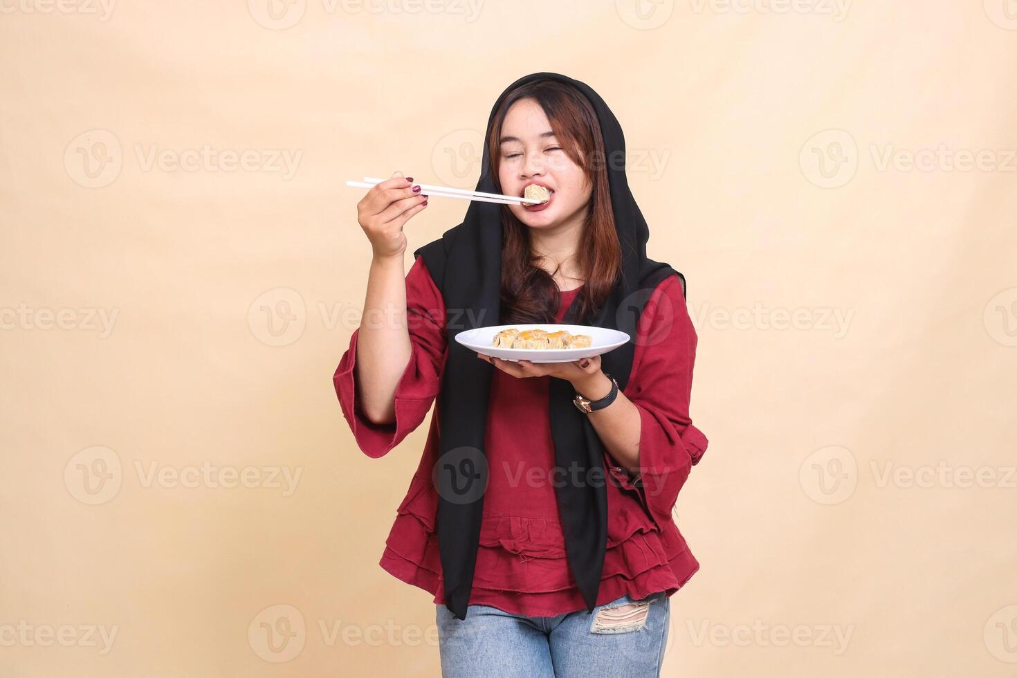 beautiful mature Asian girl wearing a red dress wearing a hijab with a delicious smile, closing her eyes, biting into dimsum using chopsticks and carrying a plate containing dimsum Chinese food photo