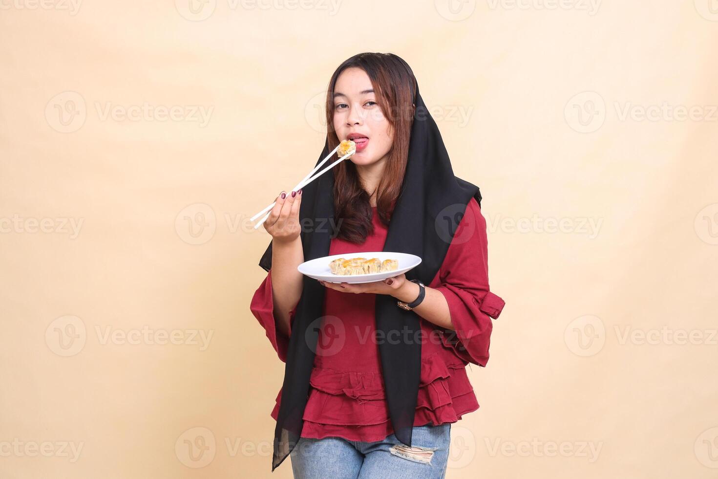 beautiful mature Asian woman in red dress wearing a hijab mockingly eats dimsum with chopsticks and brings a plate containing dimsum Chinese food to the front. used for food and culinary content photo
