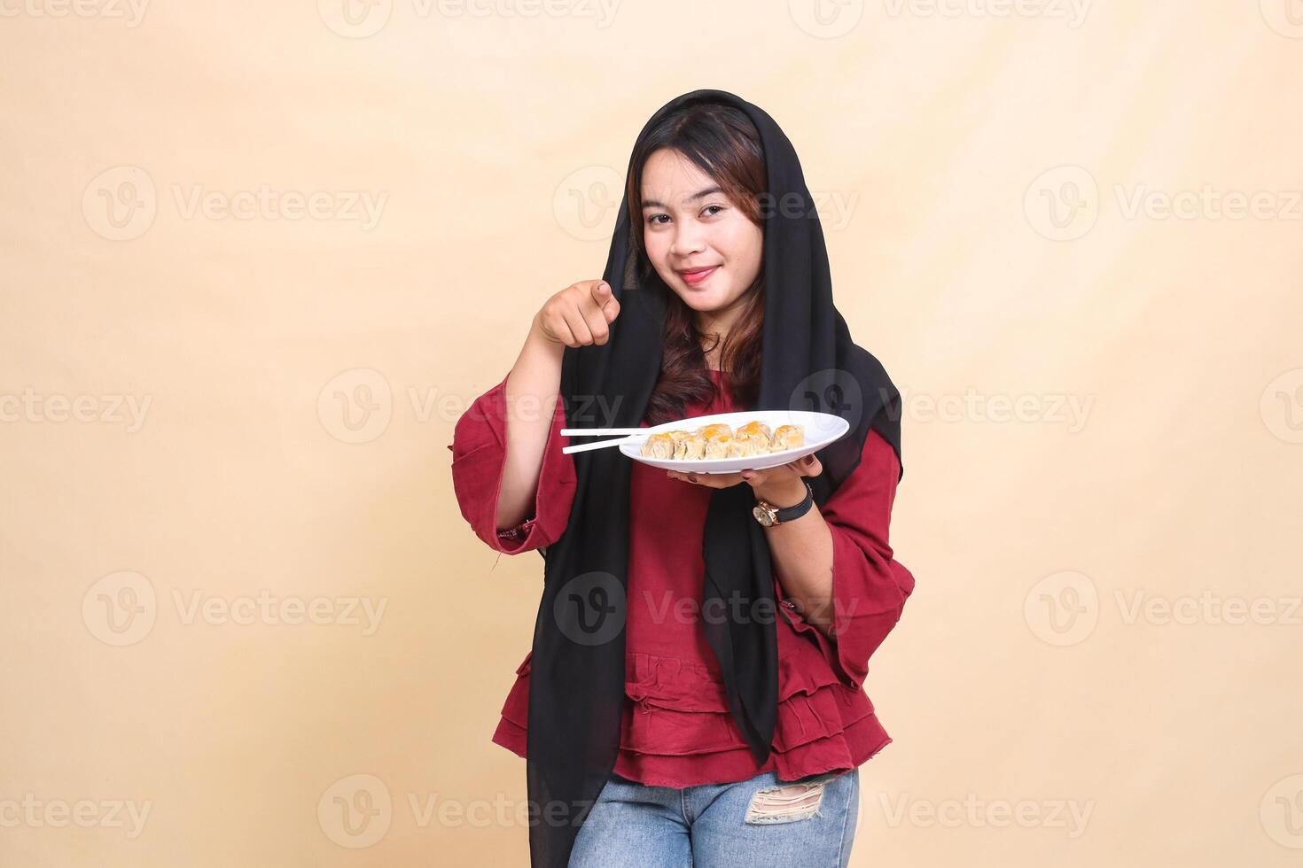 Beautiful mature indonesia woman wearing a red hijab smiling, pointing at the camera and carrying a plate containing chopsticks and dimsum Chinese food. used for food, health and lifestyle content photo