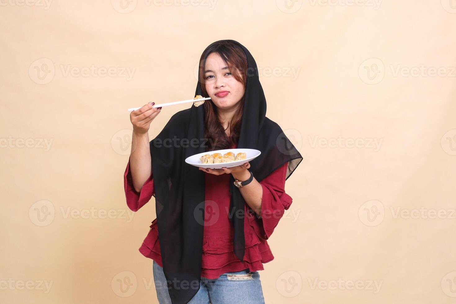 A beautiful mature indonesia woman in a red dress wearing a hijab pouting, lifting dimsum with chopsticks and carrying a plate containing dimsum Chinese food. used for health and culinary content photo