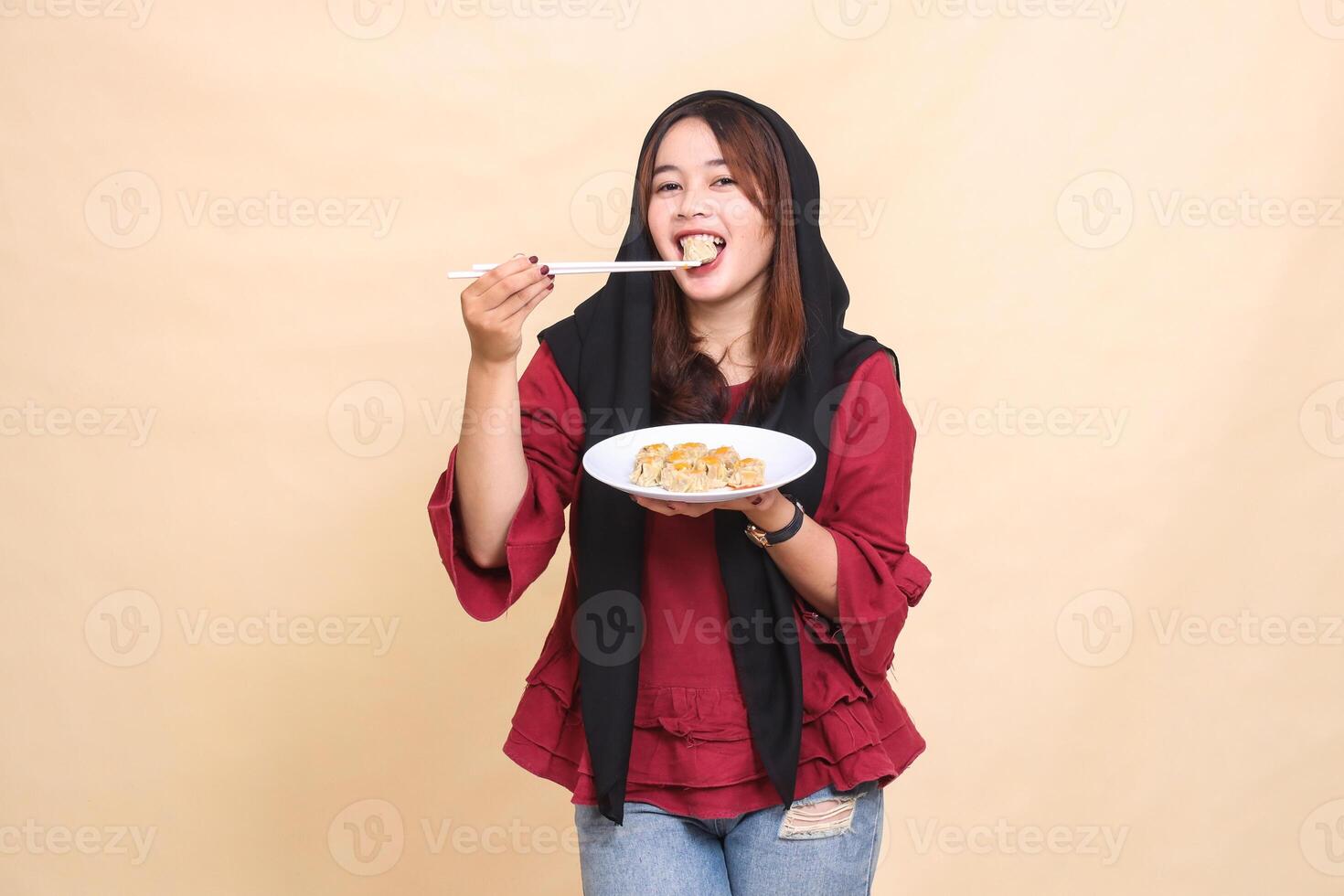 beautiful mature Asian woman wearing a red hijab, smiling, biting dimsum, using chopsticks up and carrying a plate containing dimsum Chinese food looking at the camera. used for culinary content photo