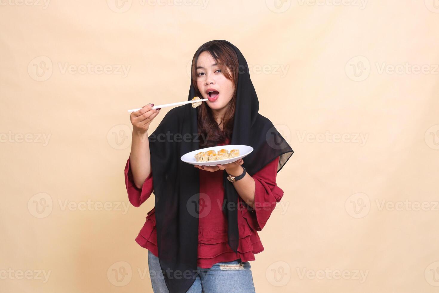 A beautiful mature Asian woman in a red dress wearing a hijab is devouring by lifting dimsum with chopsticks and carrying a plate containing dimsum Chinese food. used for food and culinary content photo