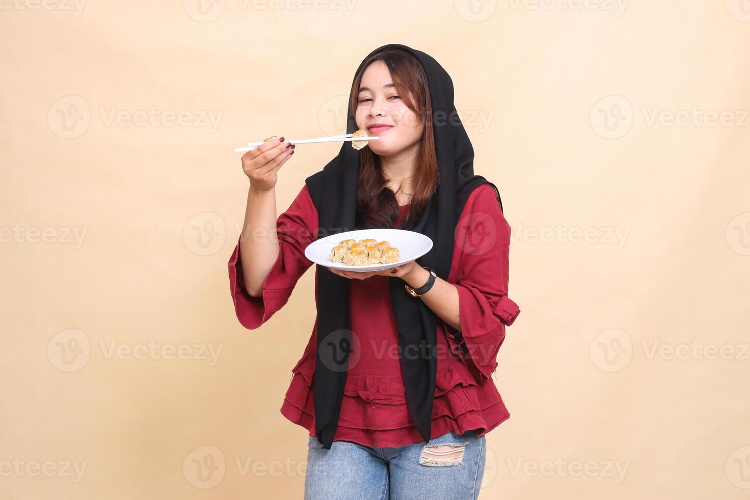 Beautiful adult indonesia girl wearing a red hijab wearing a shy smile eating dimsum with chopsticks up and carrying a plate containing dimsum Chinese food looking at the camera photo