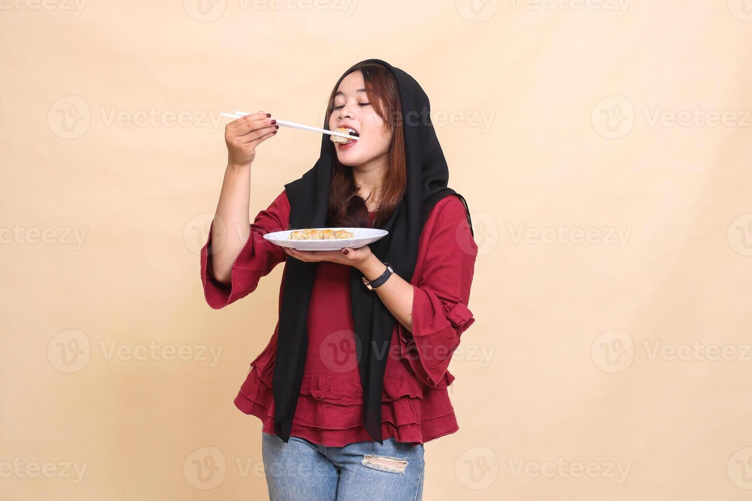 Beautiful mature indonesia woman wearing a red hijab poses, bites into dimsum with chopsticks up and carries a plate containing dimsum Chinese food. used for food, health and culinary content photo