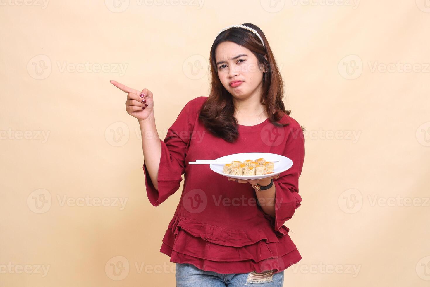 Beautiful young Asian girl in red shirt is disappointed carrying a plate containing dimsum Chinese food and chopsticks while pointing to the right. used for food, health and lifestyle content photo