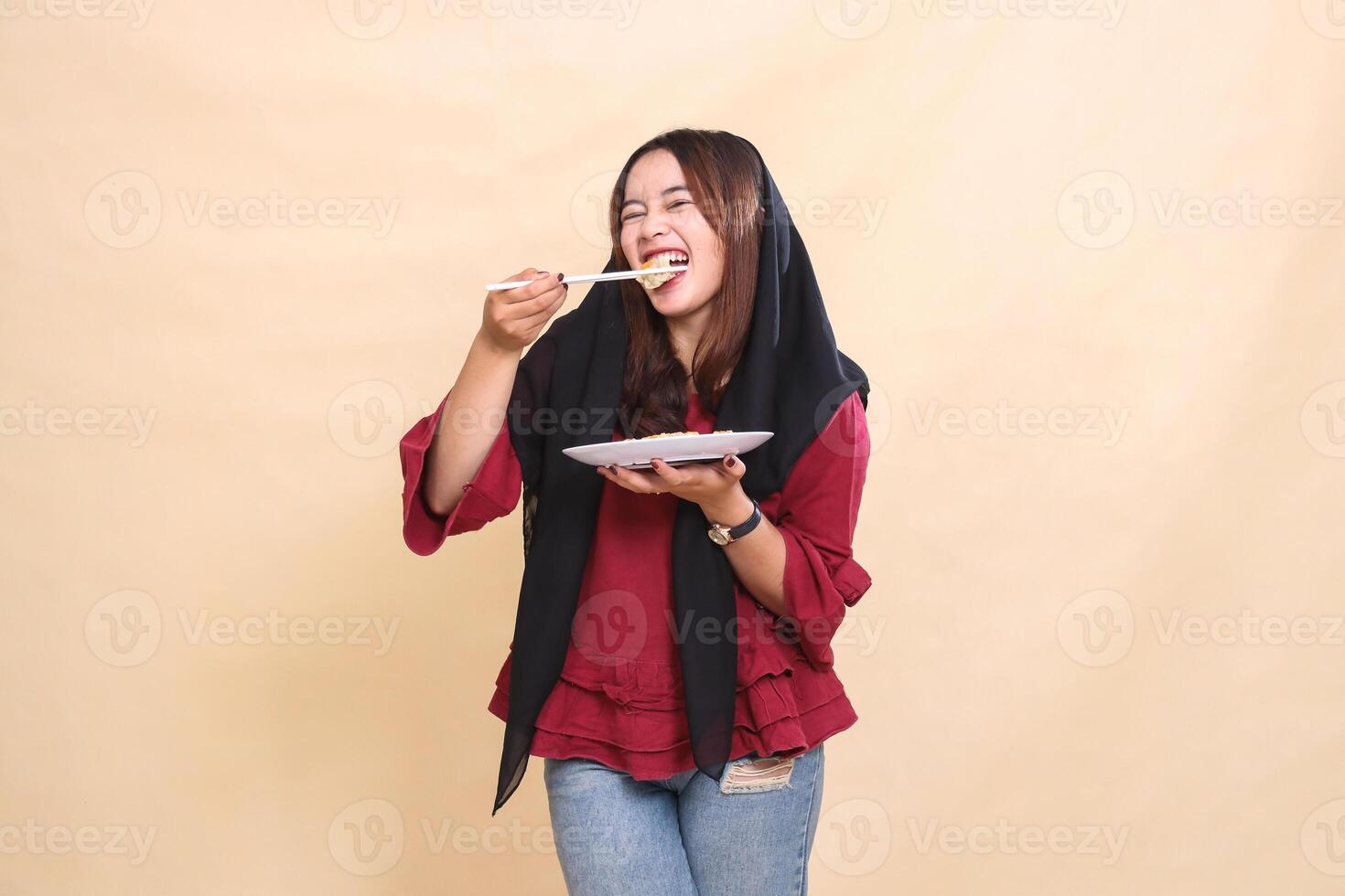 Beautiful mature indonesia woman in red shirt wearing a hijab bites dimsum with chopsticks cheerfully and carries a plate containing dimsum Chinese food candid. used for food and culinary content photo