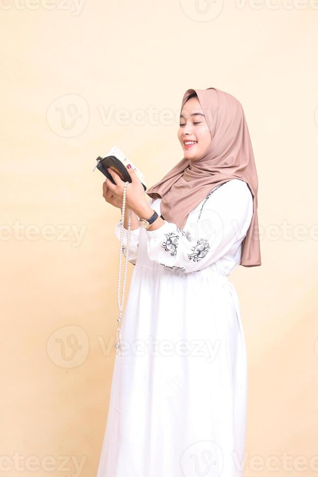 beautiful Asian Muslim adult girl in hijab to the right smiling holding prayer beads while reading the quran. for advertising, lifestyle, banners and Ramadan photo