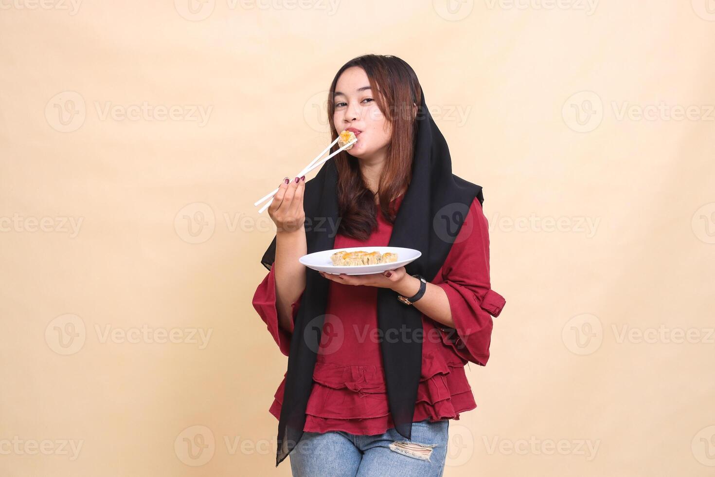 Beautiful mature Asian woman in red dress in hijab kisses dimsum with chopsticks and brings a plate containing dimsum Chinese food to the front. used for food, health and culinary content photo