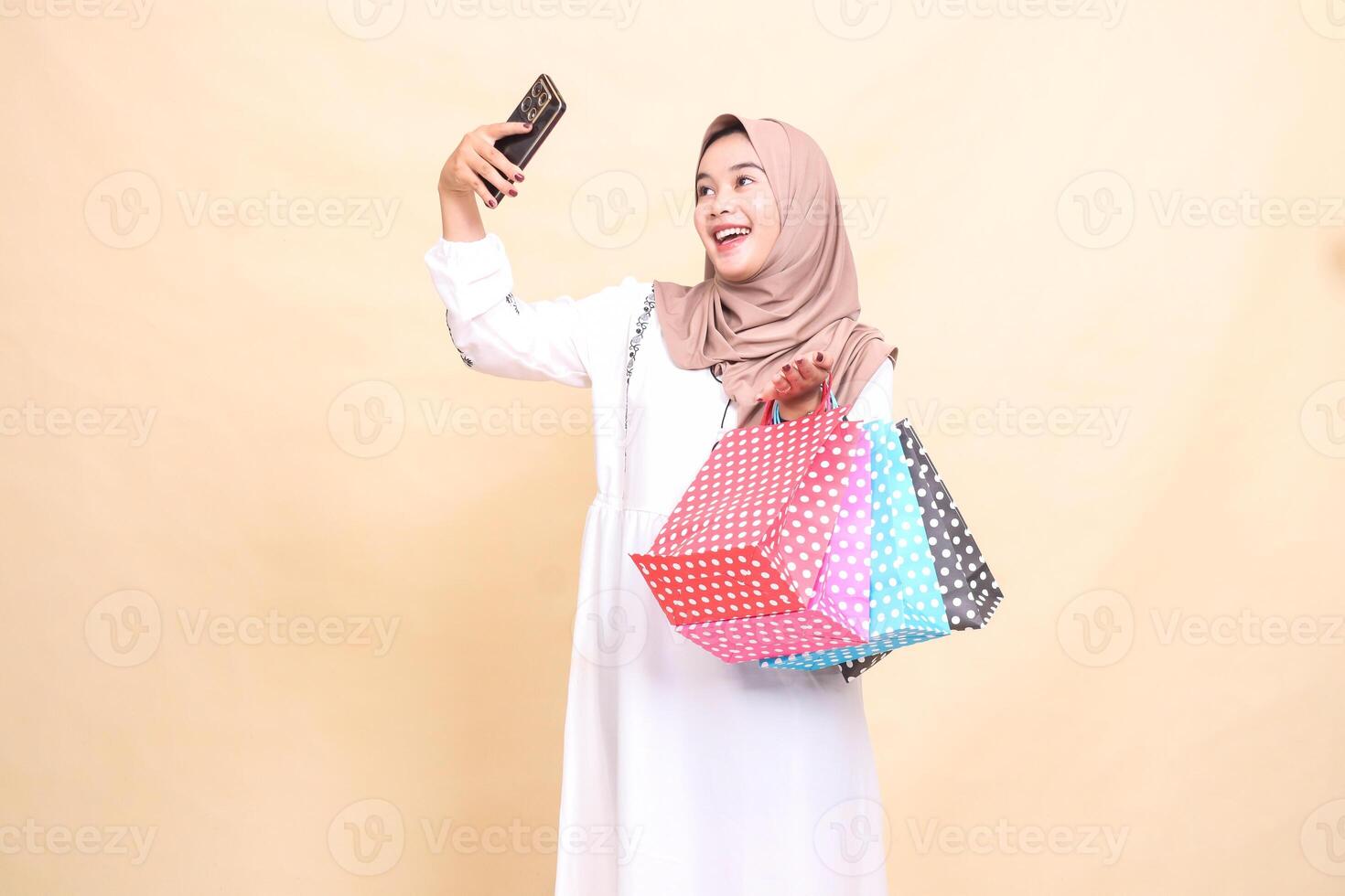 A young Asian Muslim woman wearing a cheerful hijab takes a selfie in front of a cellphone camera and carries a paper bag. for advertising, culture, Eid and Ramadan photo
