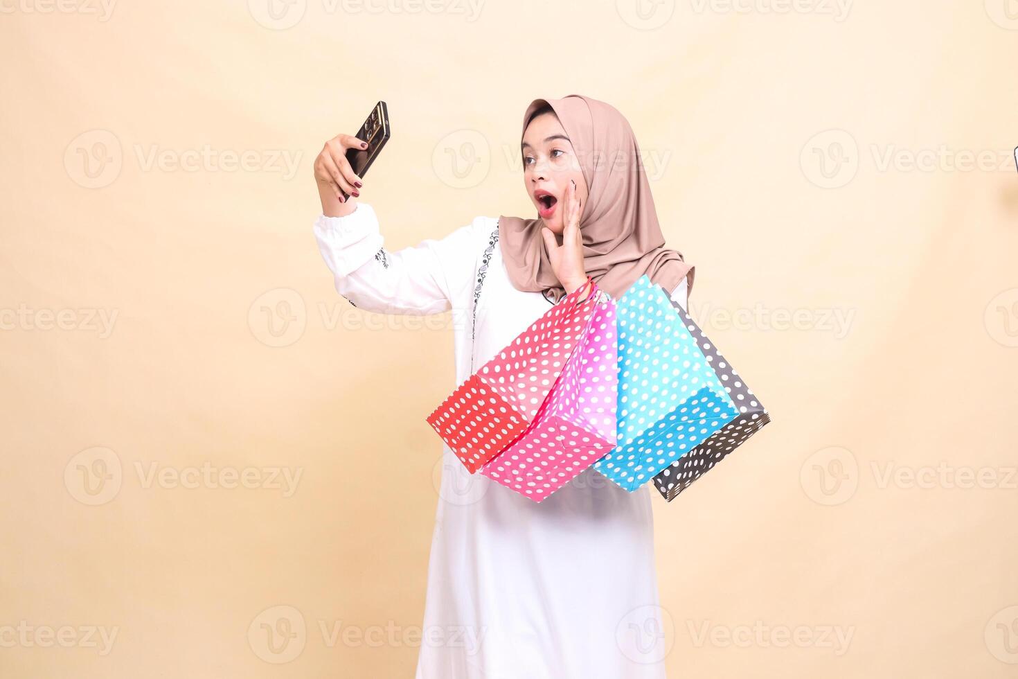 a young Asian Muslim woman wearing a hijab takes a surprised selfie greeting a cellphone camera and carrying a paperbag shopping bag. for advertising, culture, Eid and Ramadan photo