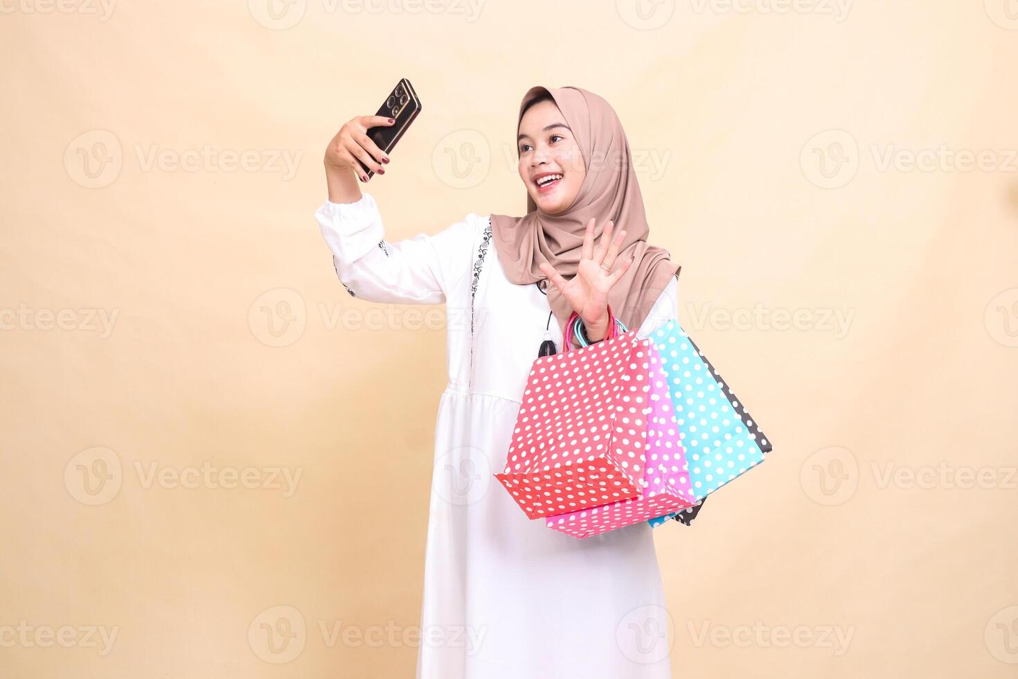a young indonesia Muslim woman wearing a cheerful hijab on a call greets in front of a cellphone camera and carries a paper bag shopping bag. for advertising, culture, Eid and Ramadan photo