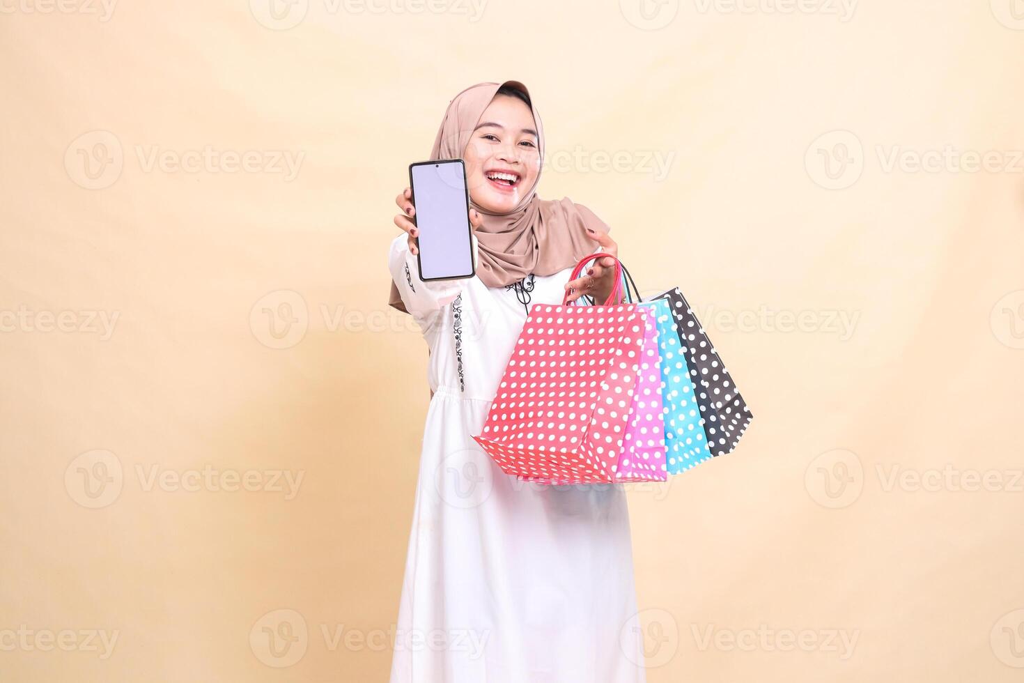 a young indonesia Muslim woman wearing a hijab laughs cheerfully, displays a cellphone screen and carries a paper bag. for advertising, technology, Eid and Ramadan photo