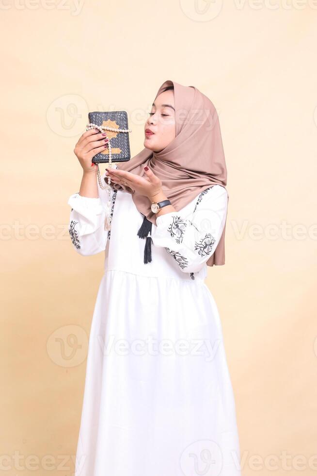 beautiful Asian Muslim mature woman in hijab candidly kissing the quran and prayer beads tied to the quran. for advertising, lifestyle, banners and Ramadan photo