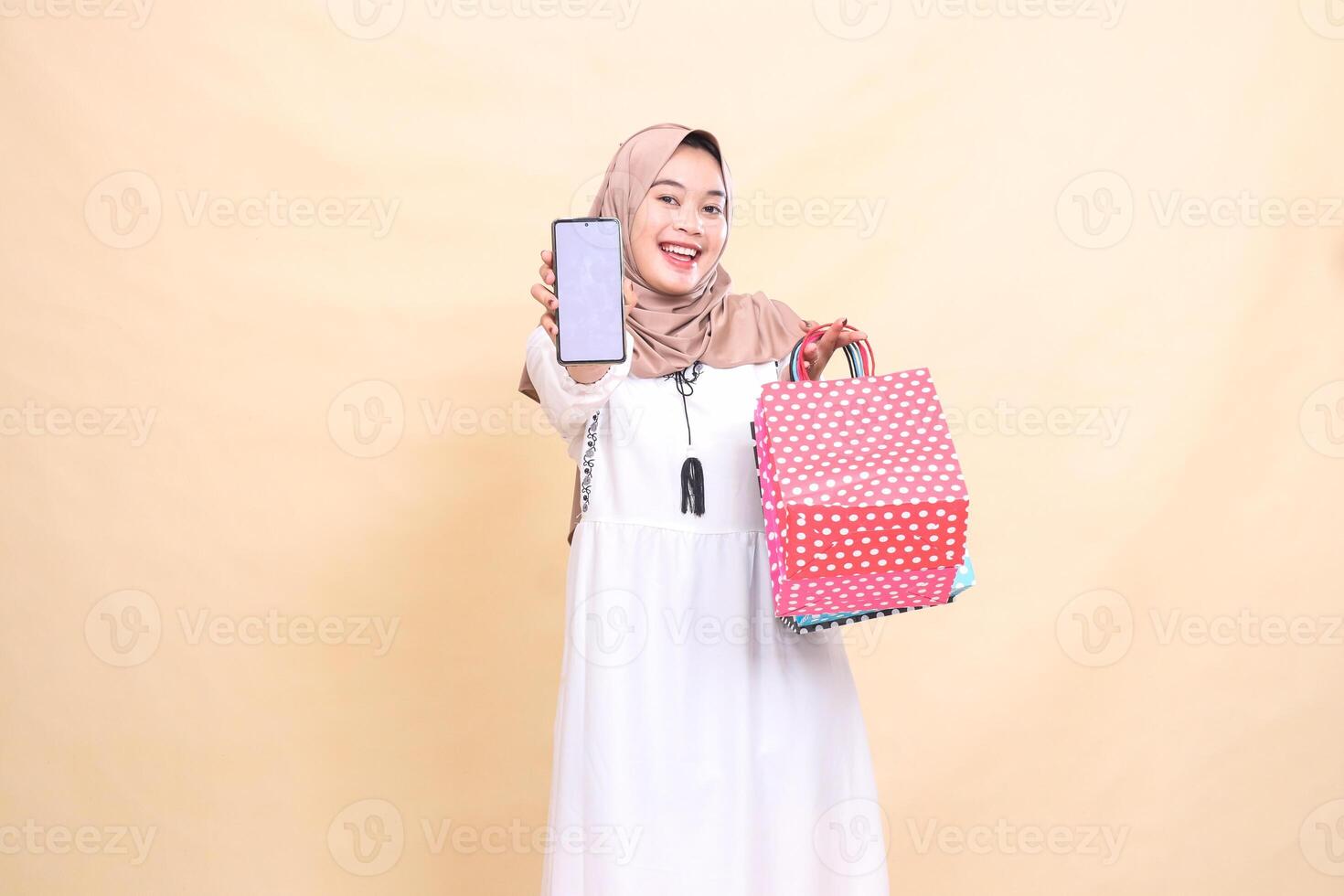 a young Asian Muslim woman in a happy hijab displays a cellphone screen and carries a paperbag shopping bag. for advertising, technology, Eid and Ramadan photo