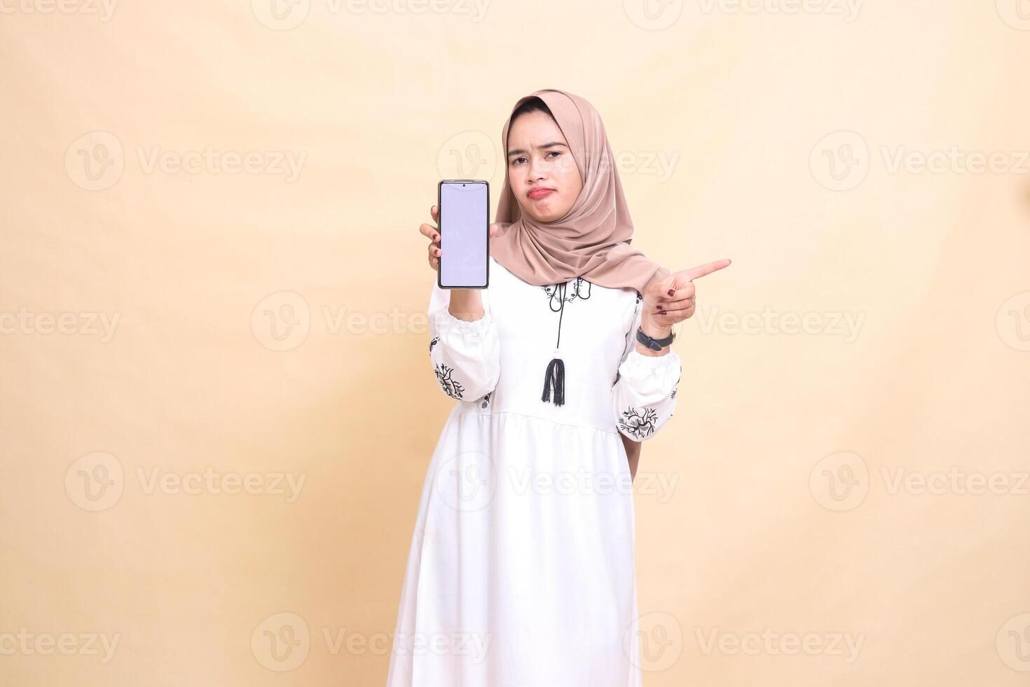 An indonesia Muslim girl wearing a hijab frowns showing a cell phone screen gadget pointing left to create sales content. for advertising, technology, Eid and Ramadan photo