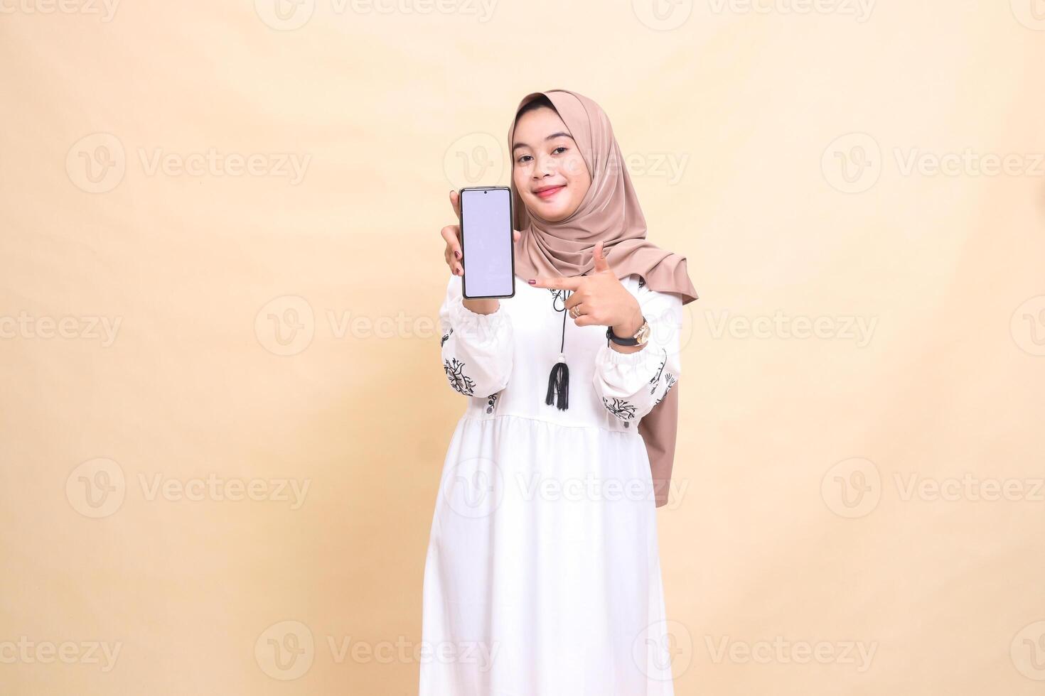 An adult Asian Muslim woman wearing a hijab smiles showing her cell phone gadget screen with a thumbs up gesture okay to create sales content. for advertising, technology, Eid and Ramadan photo