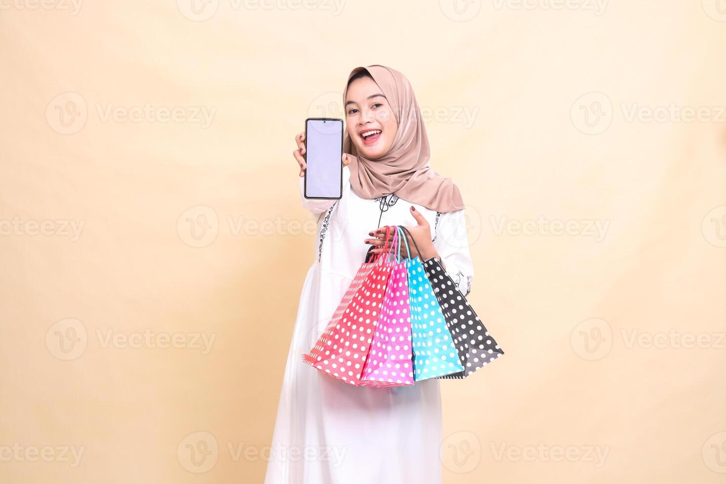 a young indonesia Muslim woman wearing a hijab is happy to display a cellphone screen and carrying a paperbag shopping bag. for advertising, technology, Eid and Ramadan photo