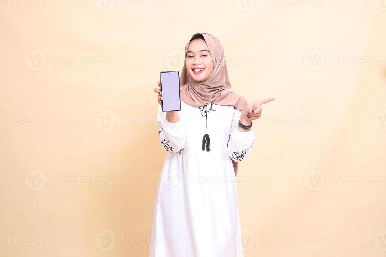 an adult indonesia Muslim woman wearing a hijab with a cheerful smile displays a cellphone screen gadget pointing left to create sales content. for advertising, technology, Eid and Ramadan photo