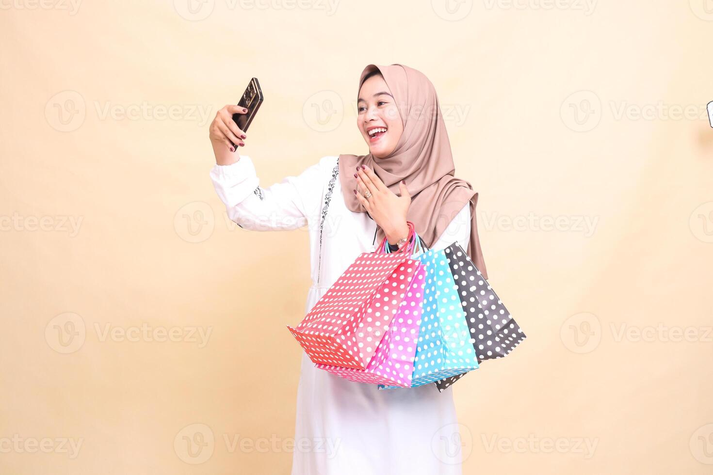 a young Asian Muslim woman wearing a cheerful hijab on a call greets her family via cellphone and carries paper bags. for advertising, culture, Eid and Ramadan photo