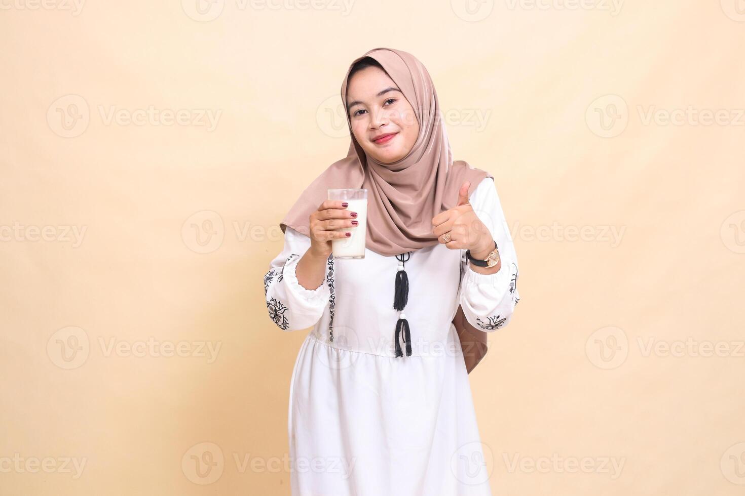 A beautiful Asian Muslim woman wearing a hijab with a smiling expression giving a thumbs up gesture carrying a glass of white milk to break the fast. used for advertising, health, Eid and Ramadan photo