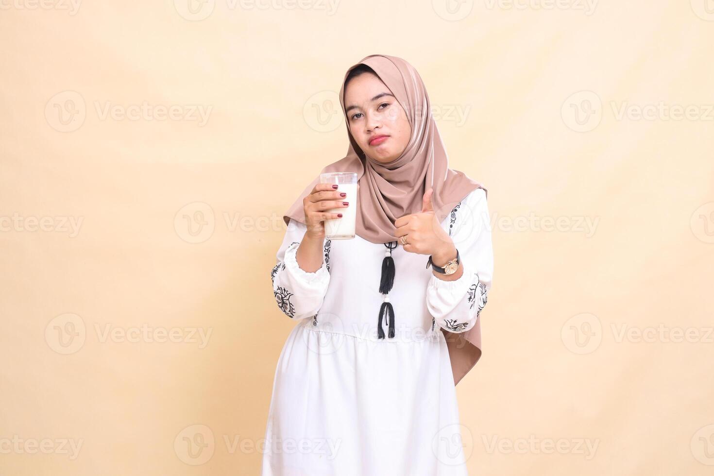 A beautiful indonesia Muslim woman wearing a hijab with a sullen expression gesturing with a thumbs up gesture carrying a glass of white milk to break the fast. used for advertising, Eid and Ramadan photo