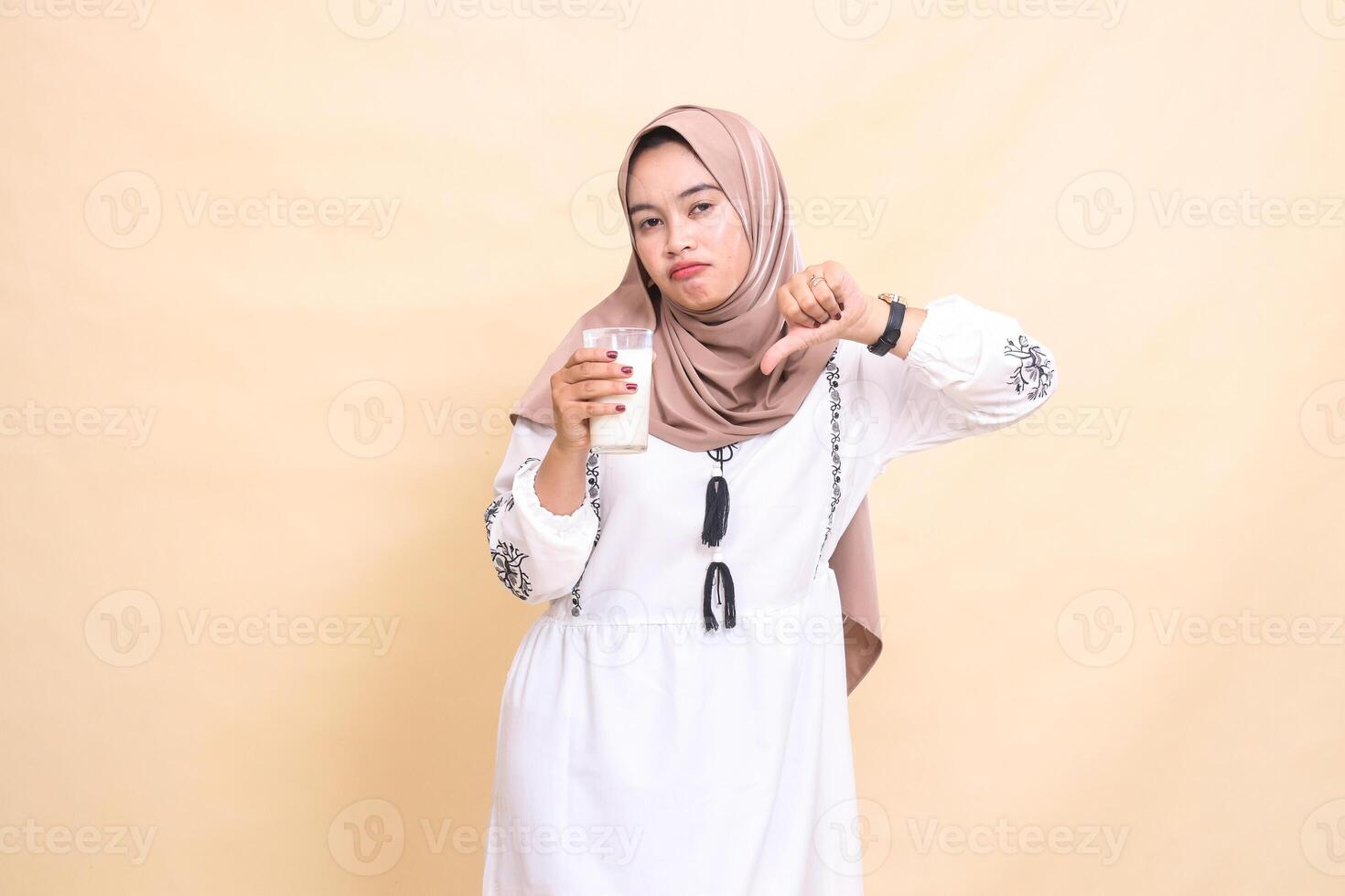 a beautiful Asian Muslim woman wearing a hijab with a sullen expression giving a bad thumbs up gesture carrying a glass of white milk to break the fast. used for advertising, health, Eid and Ramadan photo