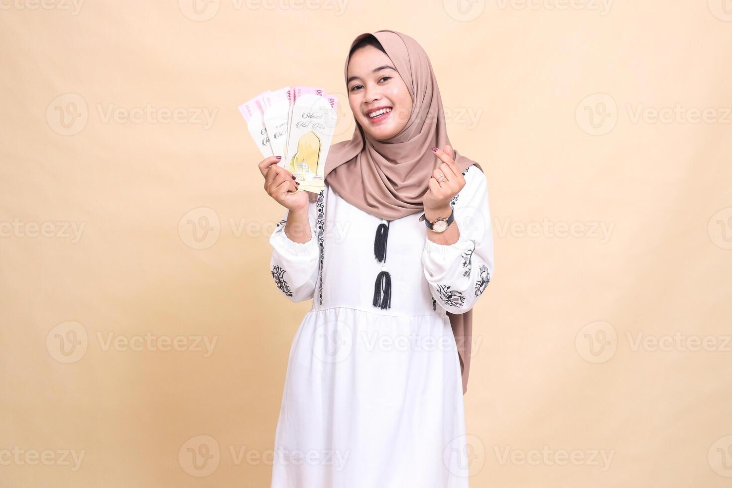 portrait of a beautiful Asian Muslim woman wearing a hijab smiling cheerfully as a gesture of love saranghaeyo holding a gift or fitrah to the right on Eid day. used for advertising, Eid and Ramadan photo