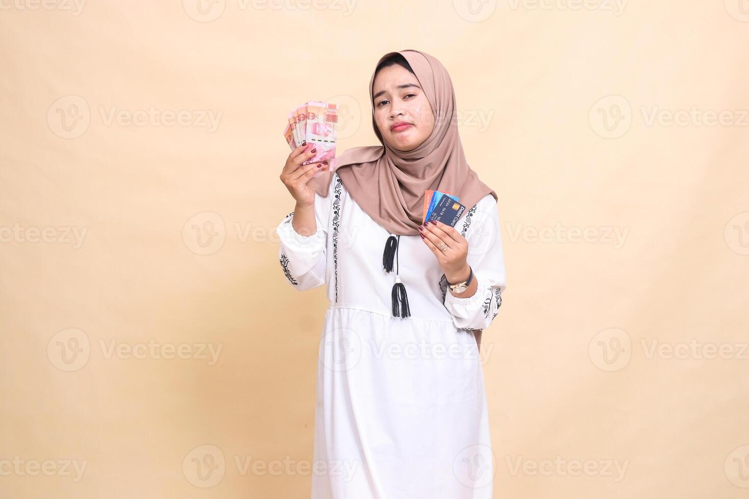 a beautiful Asian Muslim woman wearing a hijab with a disappointed expression carrying rupiah and a debit credit card for payment. used for advertising, technology, Eid and Ramadan photo
