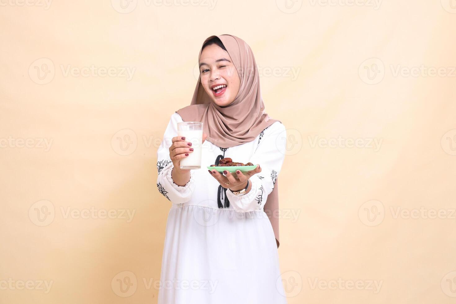 A beautiful indonesia Muslim girl wearing a hijab smiles cheerfully and winks, bringing a glass of milk and dates to the front while breaking the fast. used for advertising, health, Eid and Ramadan photo