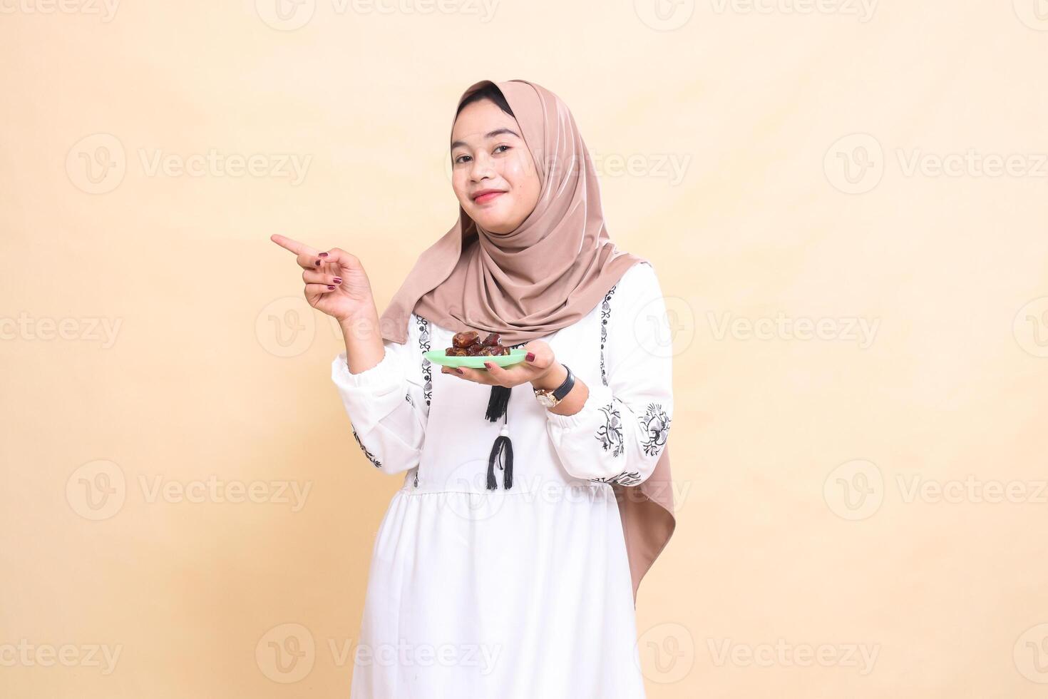 a beautiful Asian Muslim woman wearing a hijab smiles, pointing to the right and carrying dates for the sahur menu and breaking the fast. used for advertising, health, Eid and Ramadan photo
