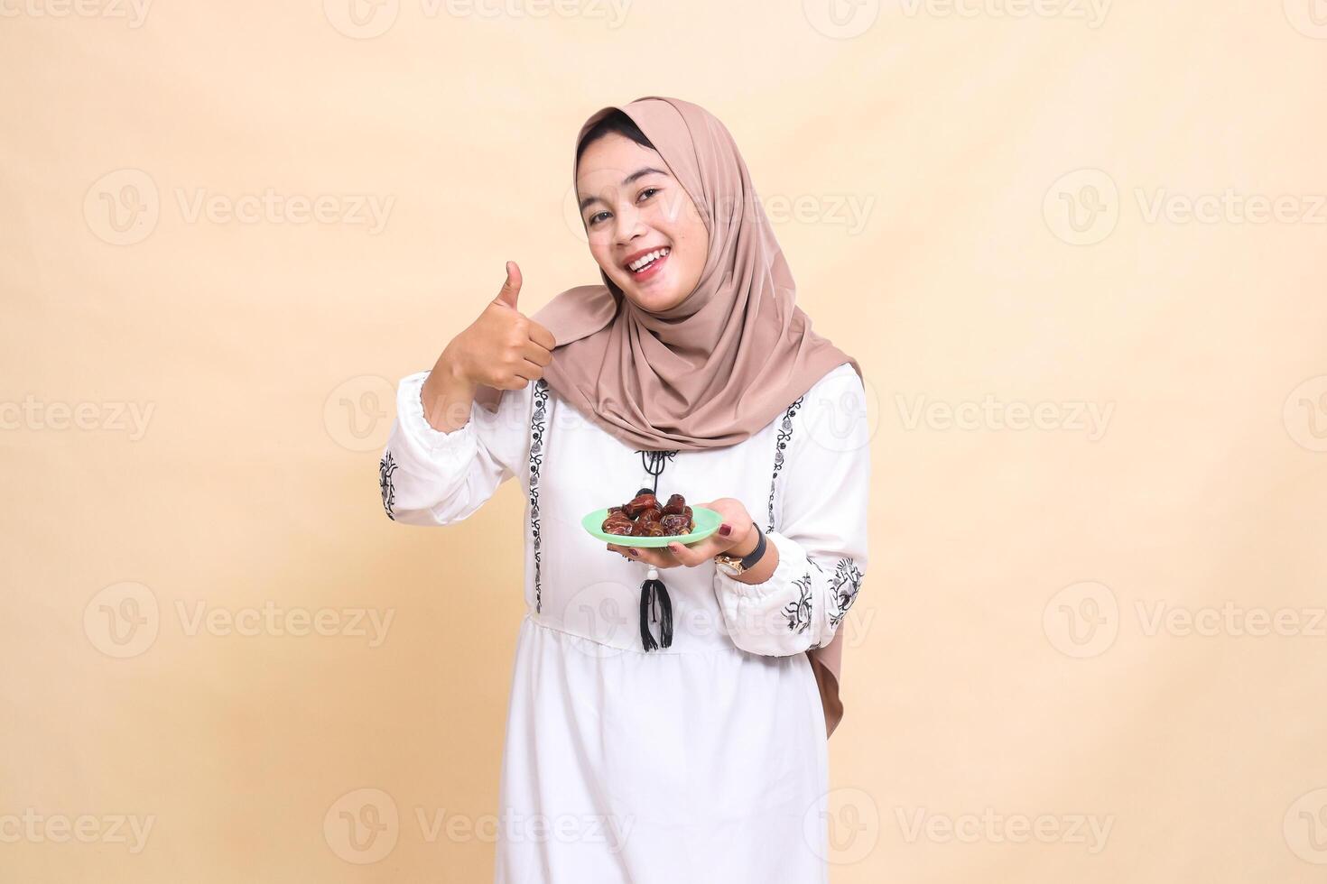 a beautiful Asian Muslim woman wearing a hijab smiles cheerfully with a thumbs up gesture okay good and brings some dates for the sahur menu and breaking the fast. used for health, Eid and Ramadan photo