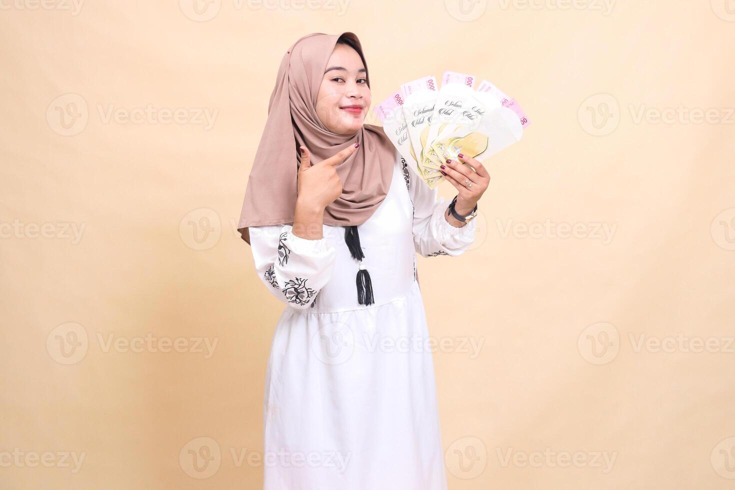 portrait of a beautiful Asian Muslim woman wearing a hijab, smiling gracefully, holding and pointing to a gift or fitrah to the left on Eid day. used for advertising, giveaways, Eid and Ramadan photo