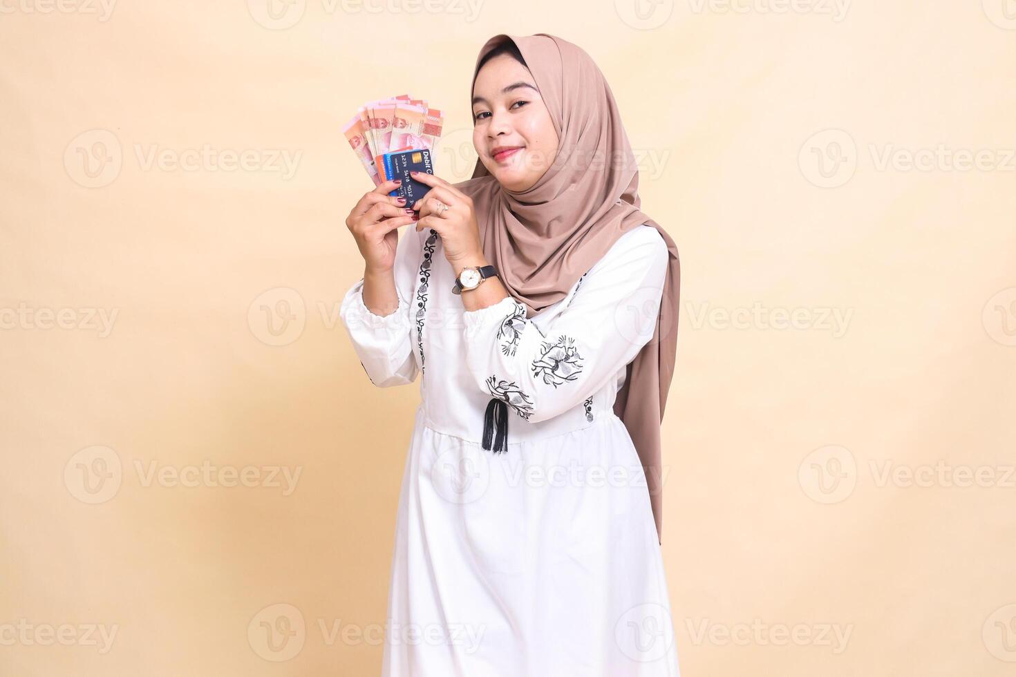 A beautiful Asian Muslim woman wearing a hijab smiles and brings to her right side rupiah notes and a debit credit card for payment. used for advertising, technology, Eid and Ramadan photo