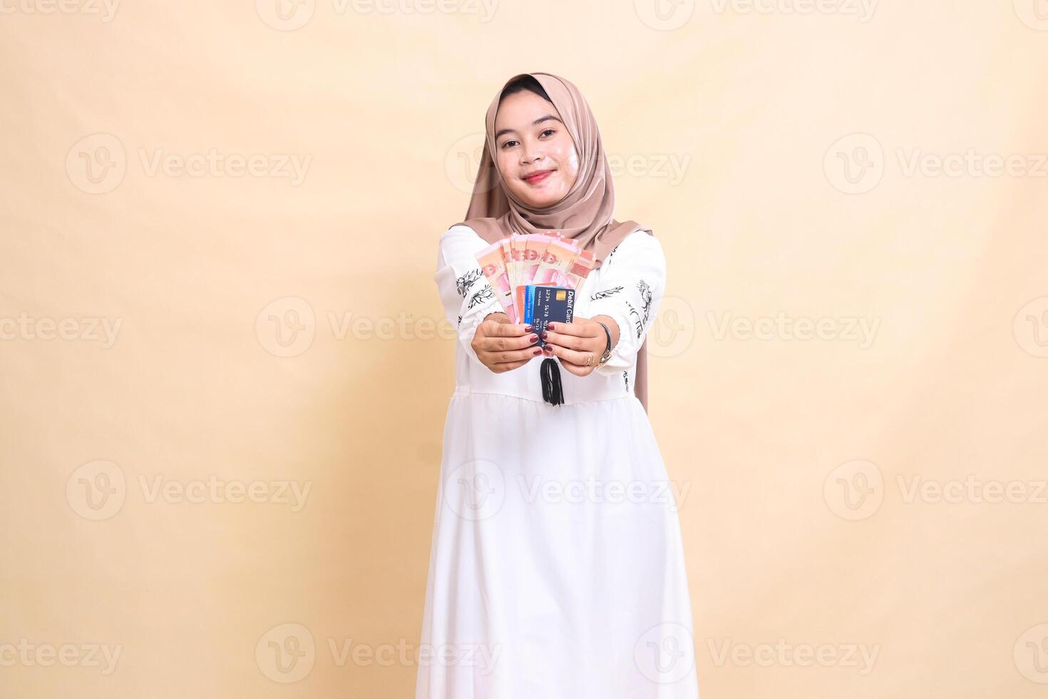 A beautiful indonesia Muslim woman wearing a hijab smiles and brings forward rupiah notes and a debit credit card for payment. used for advertising, technology, Eid and Ramadan photo