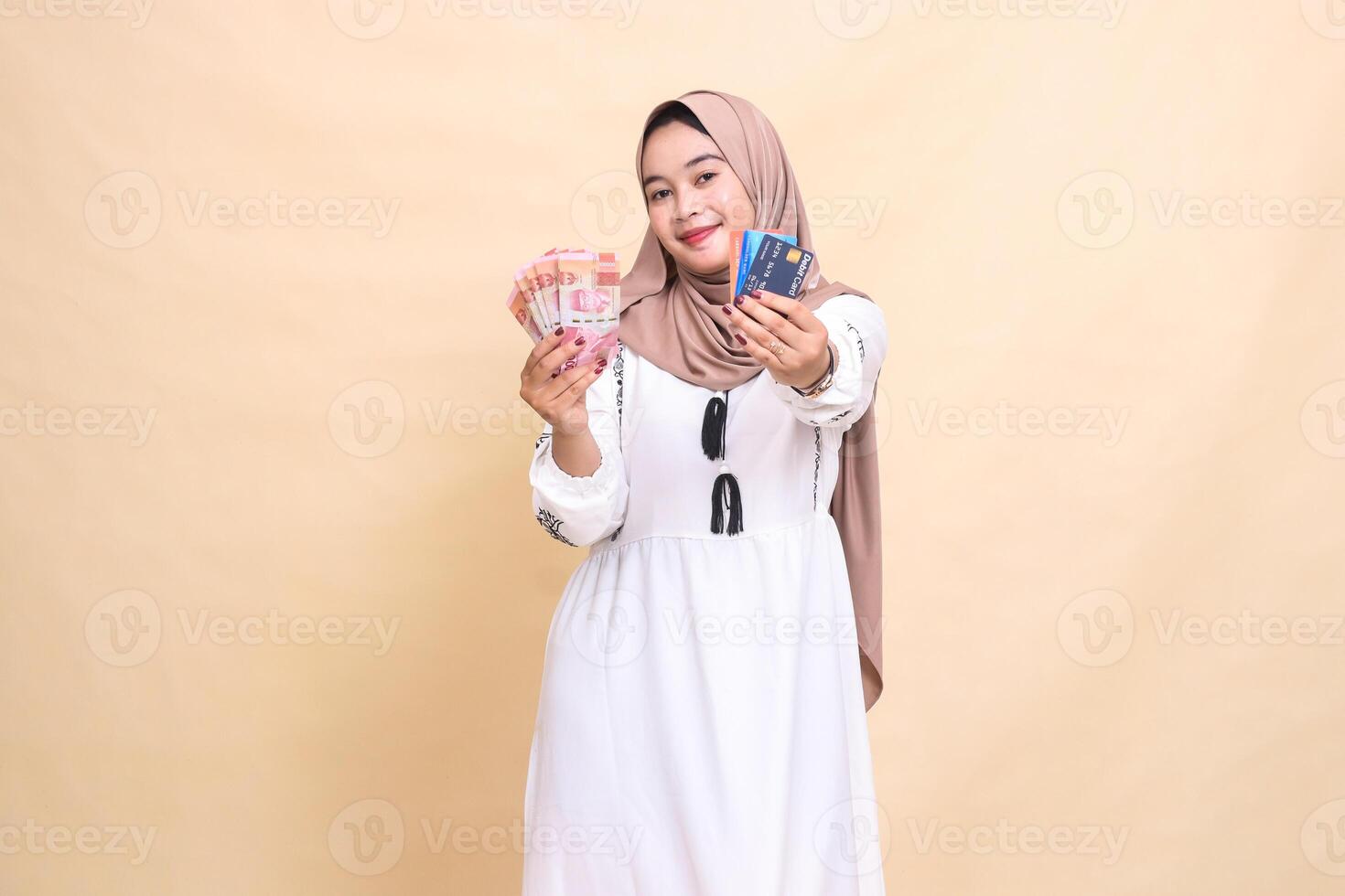 A beautiful Asian Muslim woman wearing a hijab with a smiling expression carries rupiah money and advances a debit credit card for shopping payments. used for advertising, technology, Eid and Ramadan photo