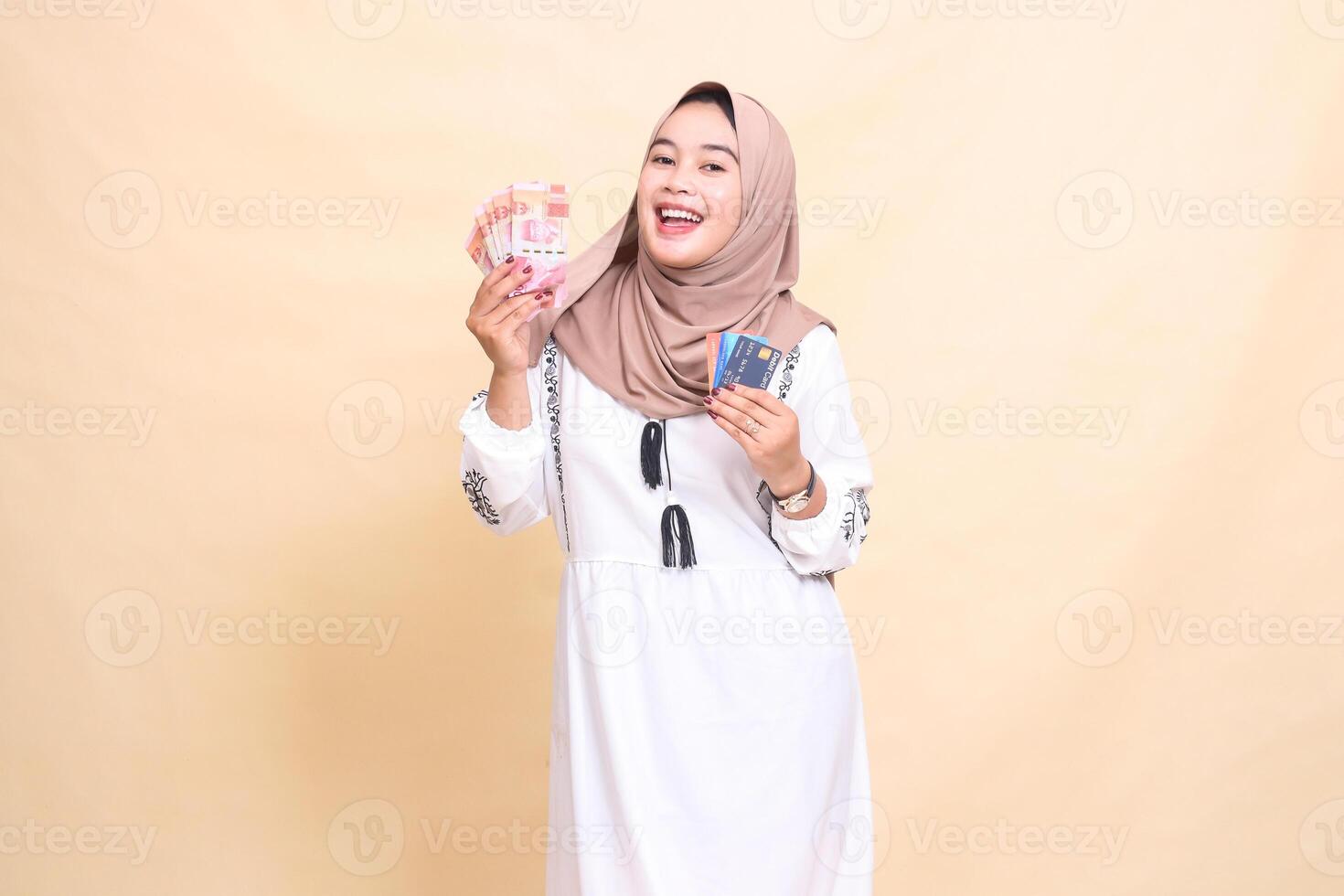 a beautiful indonesia Muslim woman wearing a hijab smiles cheerfully carrying rupiah money and a debit credit card for payment. used for advertising, technology, Eid and Ramadan photo