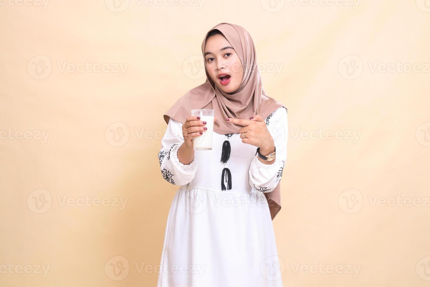 A beautiful Asian Muslim woman wearing a hijab with a shocked expression carries and points to a glass filled with white milk for eating and breaking the fast. used for advertising, health and Ramadan photo
