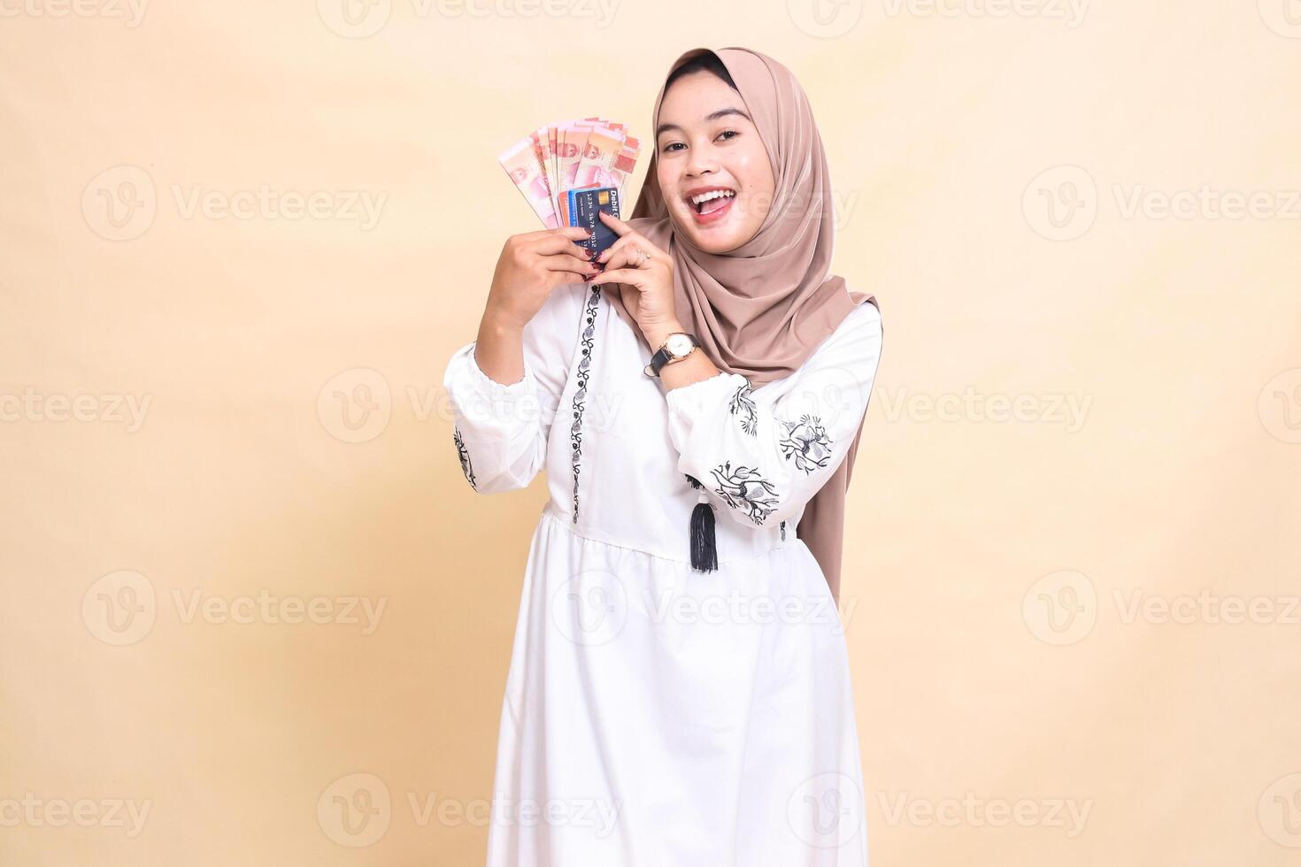 A beautiful Asian Muslim girl wearing a hijab smiles cheerfully and brings to her right side Rupiah notes and a debit credit card for payment. used for advertising, technology, Eid and Ramadan photo