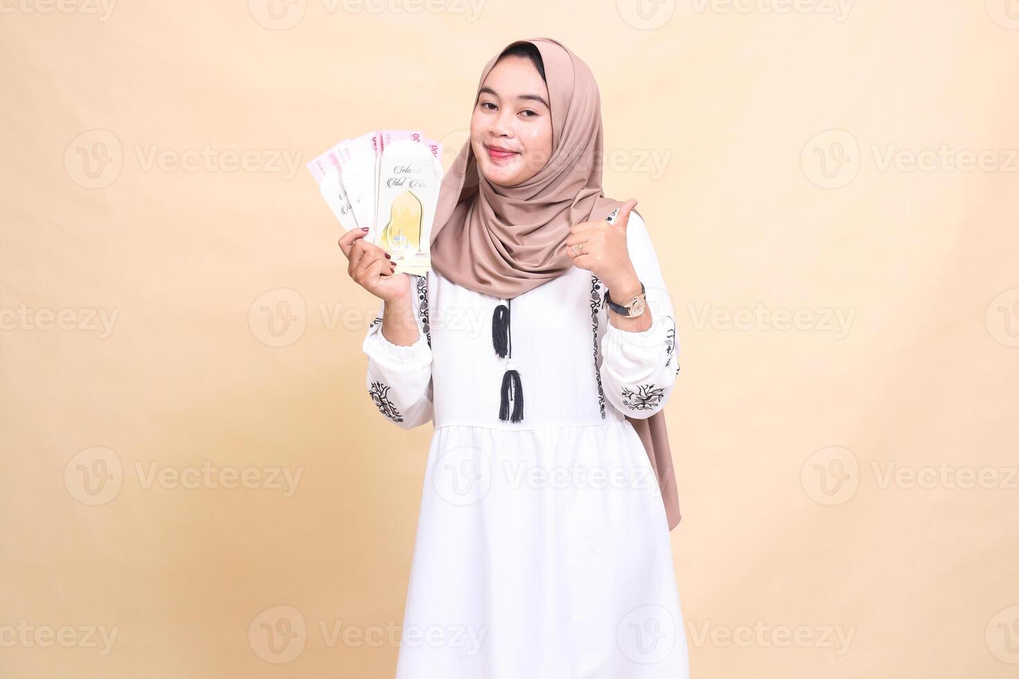 portrait of a beautiful Asian Muslim woman wearing a hijab smiling with a thumbs up gesture holding a gift or fitrah to the right on Eid day. used for advertising, giveaways, Eid and Ramadan photo
