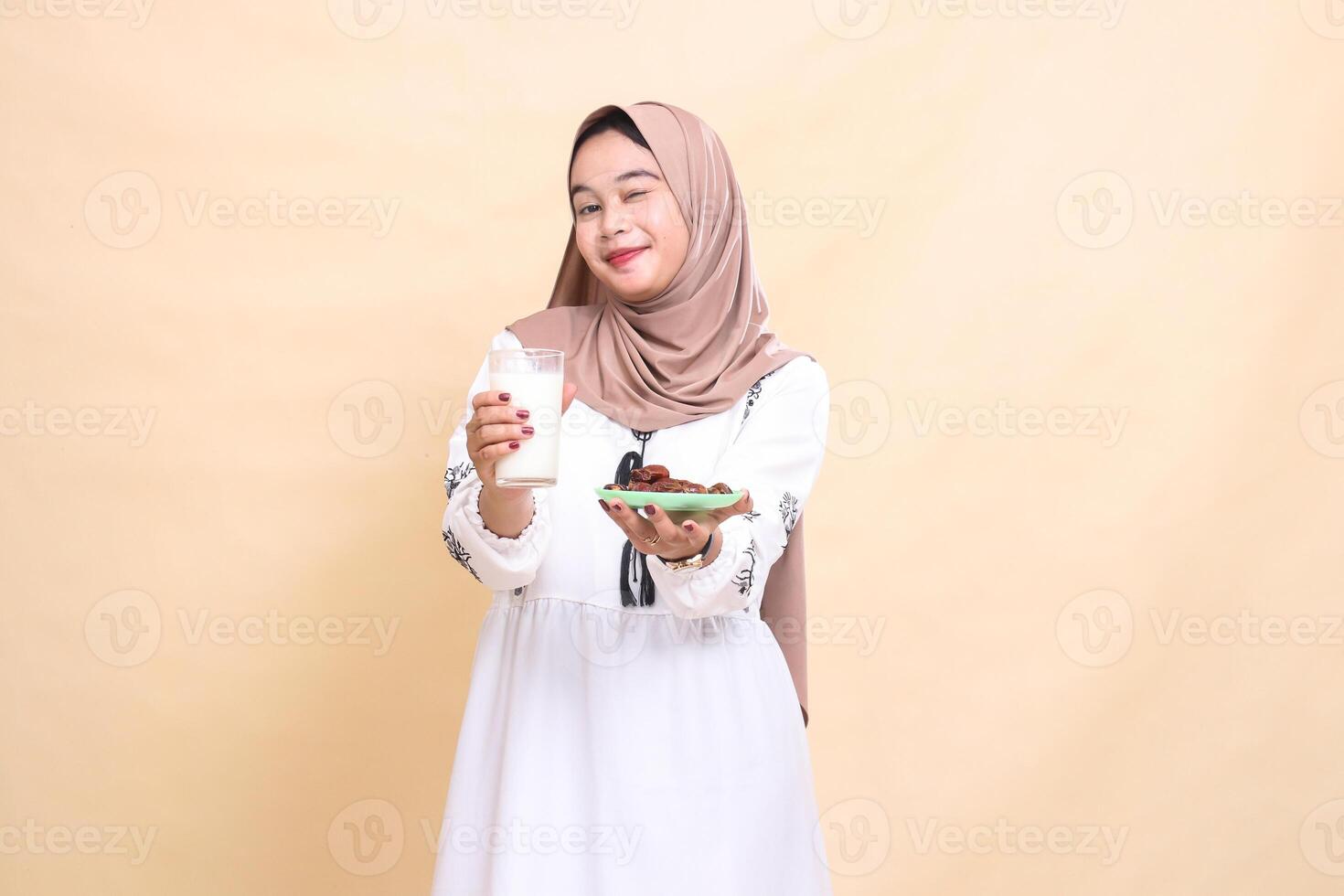 A beautiful Asian Muslim woman wearing a hijab smiles with a wink and brings a glass of milk and dates to the front while breaking the fast. used for advertising, health, Eid and Ramadan photo