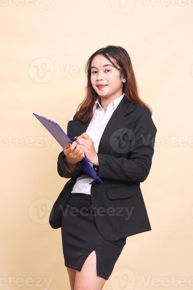 Beautiful formal Asian executive mature woman in suit smiling standing and writing pose on clipboard to camera, 3 by 4 body portrait on yellow background for commercial, vacancy and business photo