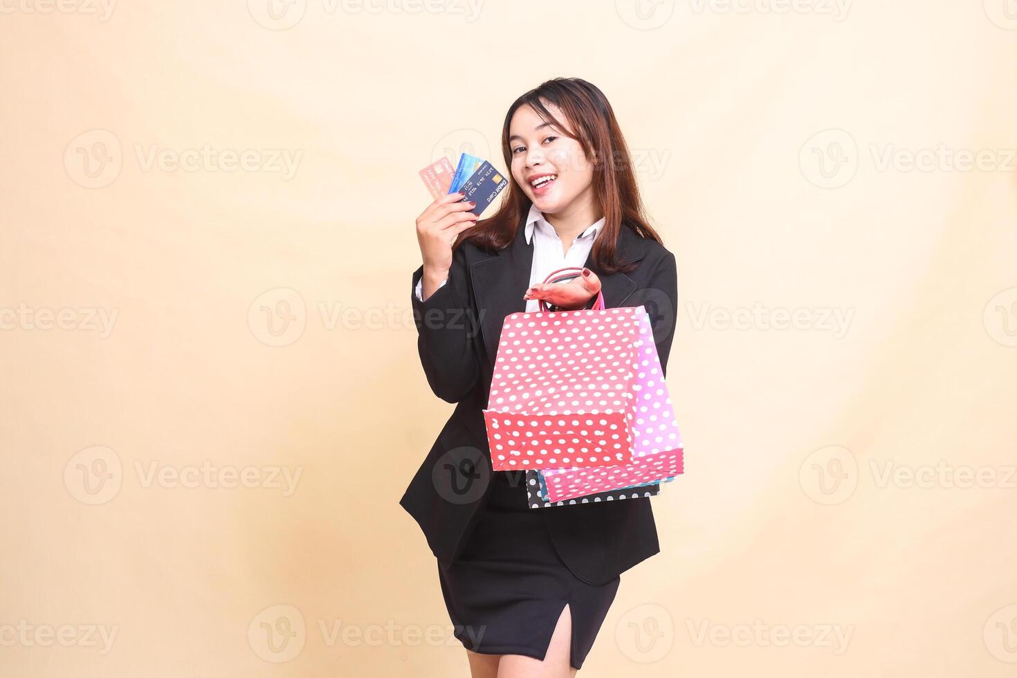 Beautiful formal Asian office woman in suit standing cheerful lifting paper bag and credit and debit cards to start paying online on yellow background for commercial, advertising and business photo