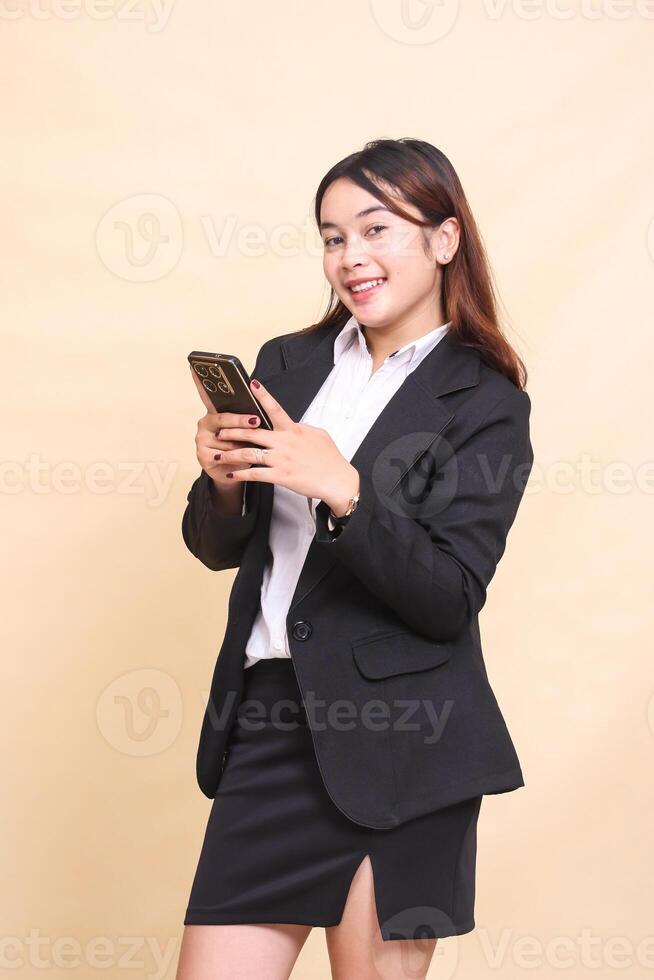 Beautiful formal Asian executive woman in suit standing cheerful smile and holding while operating online shopping gadget smartphone on yellow background for commercial, vacancy and business photo